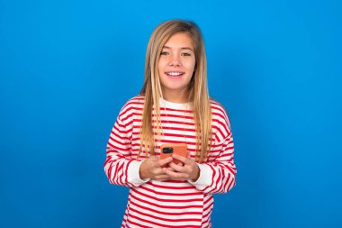 Mavi arka planda çizgili tişört giyen gülümseyen genç kız dost canlısı ve mutlu bir şekilde cep telefonuyla aynada selfie çekiyor..