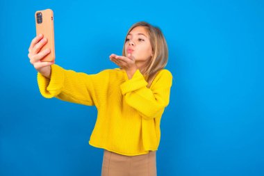 Mavi arka plan üzerine sarı süveter giyen güzel beyaz kız akıllı telefon kamerasına hava öpücüğü veriyor ve selfie çekiyor, internetten mwah gönderiyor.. 