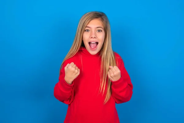 青い背景に赤いスウェットシャツを着ている美しい慎重な十代の少女は 彼女の目標と目標を達成するために幸せである喜びと彼の拳をクレンチする喜びと喜びを喜んで喜んで ポジティブな感情 — ストック写真