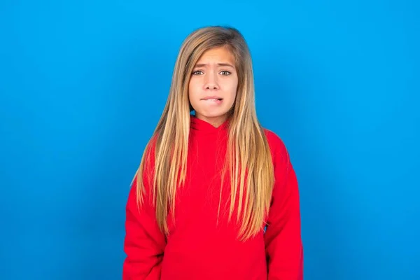青い背景に赤いスウェットシャツを着ている美しい慎重な十代の少女は緊張し 不快な表情でカメラを見ている恐ろしい唇を噛んで — ストック写真