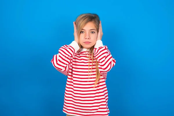 沮丧的少女穿着条纹衬衫 双手插在蓝色背景的耳朵上 不愿听摇滚乐 噪音或嘈杂的音乐 — 图库照片