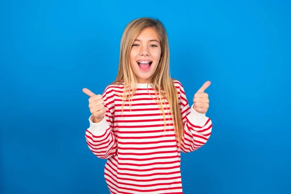 楽観的な10代の少女は ポジティブな感情で親指を示す青い背景の上にストライプシャツを着ています 品質と推奨コンセプト — ストック写真
