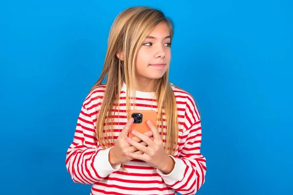 青い背景にストライプのシャツを着た10代の少女の肖像画は 夢のような外観で スマートフォンを保持しながら考えています メッセージを書くために試みる — ストック写真