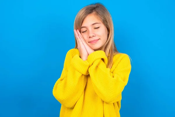 青いスタジオの背景に黄色いセーターを着用しているコーカサスティーンガールは 押された手のひらに夢を閉じ 何かについて楽しい笑顔を持っています — ストック写真