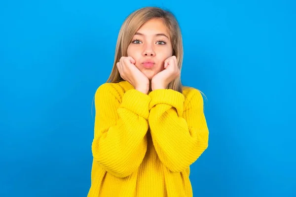 青いスタジオの背景に黄色のセーターを着て美しい白人十代の少女は驚いた表情で手を保ち 唇を折りたたみ 面白いグリムを作ります — ストック写真