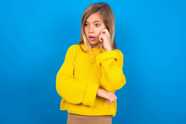 青いスタジオの背景に黄色のセーターを着用している驚くほど慎重な十代の少女は 口を開いたと驚くほど外見します — ストック写真