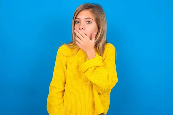 青いスタジオの背景に黄色いセーターを着用しているブロンドの慎重な十代の少女は 口をカバーし カメラで不思議に見える 予期しない噂を信じることができません — ストック写真