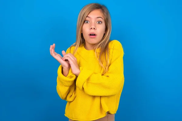 青いスタジオの背景の上に黄色のセーターを着用している驚くほどの慎重な十代の少女は 手のひらをこすり 不信感を持ってカメラを見つめます — ストック写真