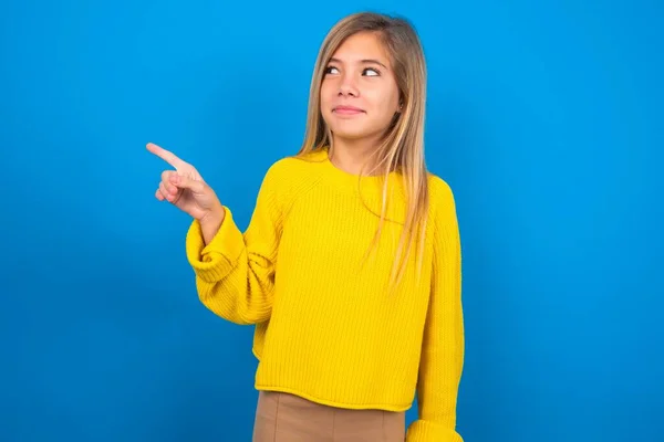 コピースペースで青いスタジオのバックグラウンドポイント上の黄色いセーターを着て魅力的な慎重な十代の少女は 何かを宣伝します 最高の価格をアドバイス — ストック写真