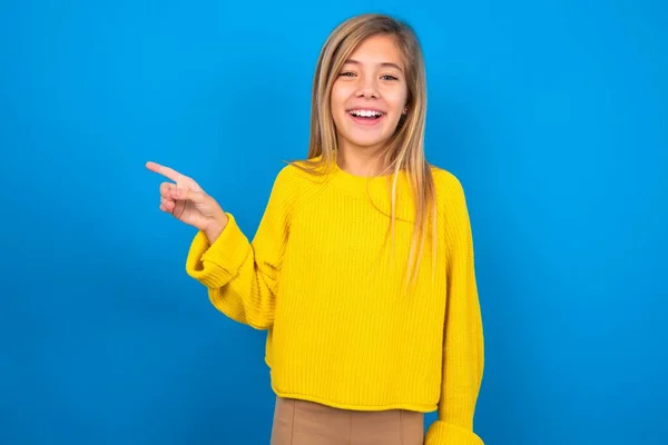 青いスタジオの背景に黄色のセーターを着ている美しい白人十代の少女は幸せに空白のスペースで離れて指摘し 良いニュースや予想外の販売に興奮ショッピング割引オファーを示しています — ストック写真