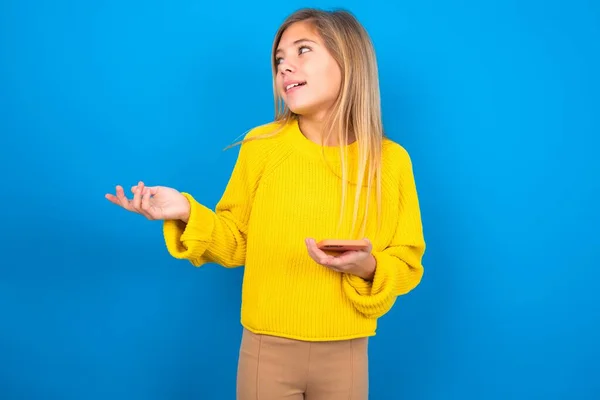 青いスタジオの背景に黄色いセーターを着用している幸せな白人十代の少女はパームを上げ 携帯電話はテキストメッセージやビデオ通話のための高速インターネットを使用しています — ストック写真