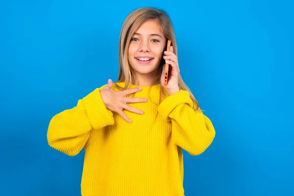 笑顔の慎重な十代の少女は 携帯電話を介して黄色いセーターのトークを着用し 青い背景で楽しい素晴らしい会話を楽しんでいます — ストック写真