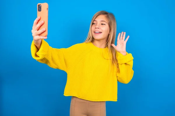 Blonde在蓝色工作室背景下穿着黄色毛衣的年轻女孩手持现代手机 并以打招呼的姿势做着视频呼叫波手掌 — 图库照片