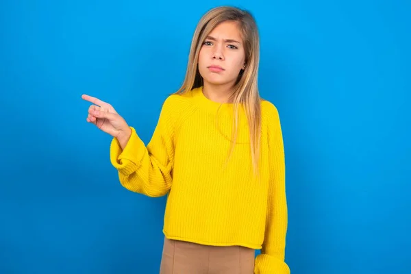 青いスタジオの背景に黄色いセーターを着用している深刻な慎重な十代の少女は コピースペースの向こうのポイントは 何か不快なことを示しています この広告を見てください ビッグプライスコンセプト — ストック写真