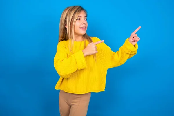 青いスタジオの背景に黄色いセーターを身に着けている美しい慎重な十代の少女の笑顔は指を指示することに興奮しました — ストック写真