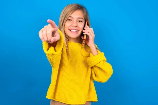 青いスタジオの背景に黄色いセーターを着用するポジティブな慎重な十代の少女は カメラで直接電話会話の笑顔を広く楽しんでいることを示しています — ストック写真