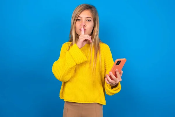 ブロンドの慎重な十代の少女は 現代のガジェットを保持している青いスタジオの背景に黄色のセーターを着用し 秘密を伝えることはありません — ストック写真