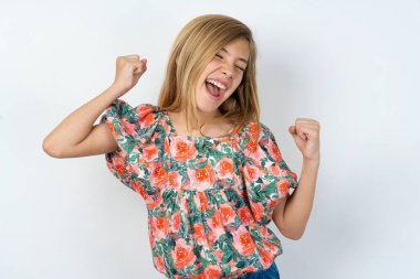 Ecstatic beautiful caucasian teen girl wearing flowered blouse over white wall shout loud yeah fist up raise win lottery clipart