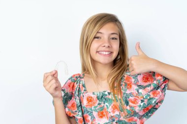 Beyaz duvarın üzerinde çiçekli bluz giyen güzel beyaz bir kız görünmez bir diş teli tutuyor ve baş parmağını kaldırıyor. Bu yeni tedaviyi öneriyor. Diş sağlığı konsepti.