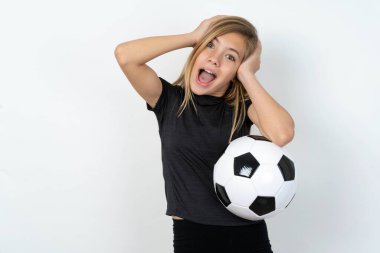Neşeli, neşe dolu, spor giysili, beyaz duvarda futbol topu tutan genç kız harika haberler aldıktan sonra ellerini havaya kaldırarak tepki veriyor..