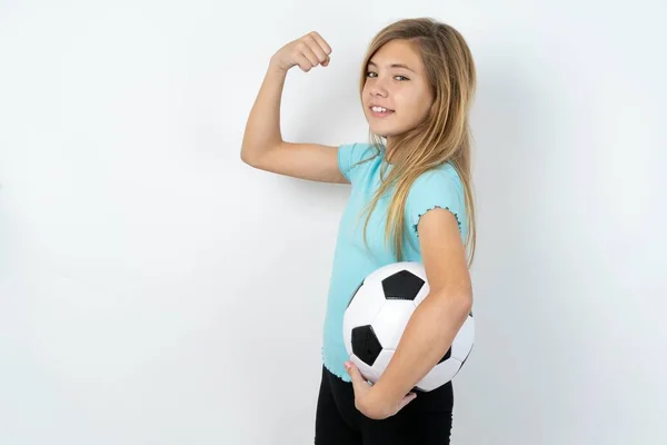 筋肉を示す白い壁の上にサッカーボールを保持するスポーツウェアを身に着けている強力な陽気な慎重な十代の少女の肖像画 — ストック写真
