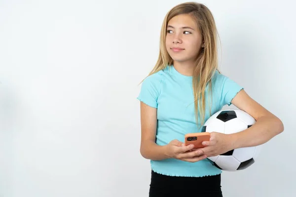 白い壁の上にサッカーボールを保持するスポーツウェアを身に着けている白人の十代の少女は電話の手を読みます 良い若者のニュースは空のスペースの広告を見ます — ストック写真