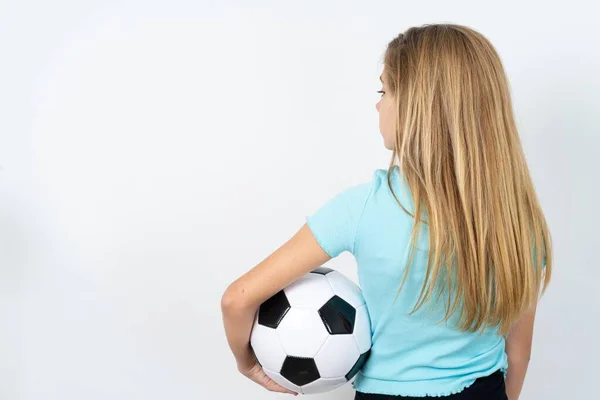白い壁の上にサッカーボールを保持するスポーツウェアを身に着けている白人十代の少女の背面の景色 スタジオショット — ストック写真