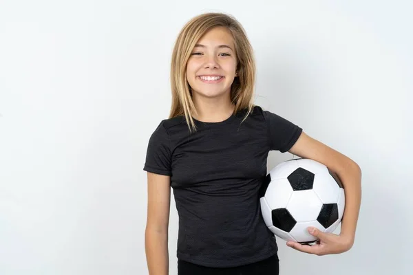 素敵な笑顔と喜びの表情で白い壁の上にサッカーボールを保持するスポーツウェアを着ているティーンガール ポジティブな感情コンセプト — ストック写真