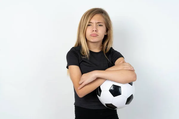 白い壁の上にサッカーボールを保持するスポーツウェアを身に着けている不満の十代の少女は額の下からカメラで悲惨な表情で 不幸な悲しみを作ります — ストック写真
