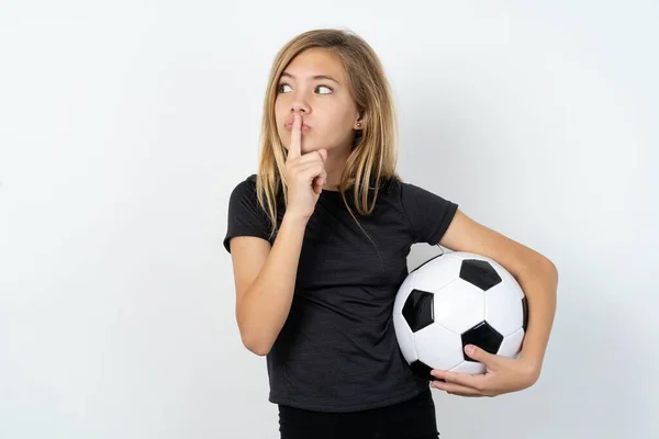 身穿运动服的少女拿着足球越过白墙 沉默的手势让食指紧闭嘴唇 做着沉默的手势 要求不要与他人分享秘密 — 图库照片