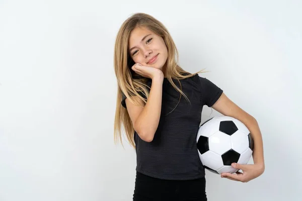 白い壁の上にサッカーボールを保持するスポーツウェアを身に着けている深刻な美しい白人少女を魅了することは カメラで優しく顔の笑顔の近くに手を保ちます — ストック写真
