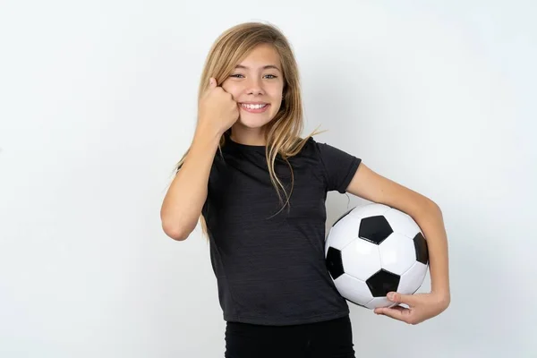 ホワイトウォールの上にサッカーボールを保持するスポーツウェアを着用ハッピーティーンガールは 頬の笑顔に拳を保ち 良い気分で肯定的な表現を持っています — ストック写真