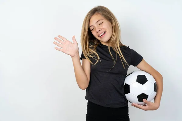 ホワイトウォールの上にサッカーボールを保持するスポーツウェアを身に着けている大喜びの成功したティーンガールは 友人によって楽しませられる喜びで目を閉じます — ストック写真