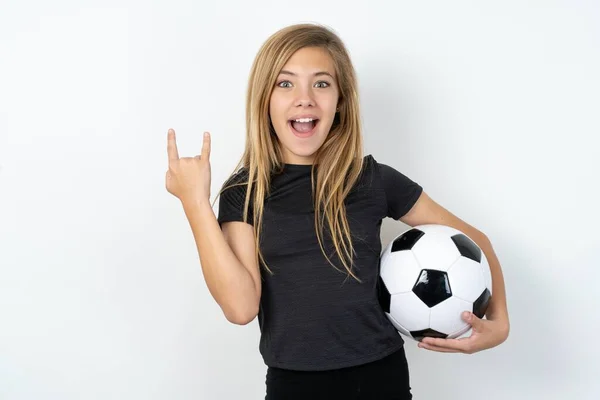 身穿运动服的少女拿着足球冲过白墙 使摇滚标志看起来自信而快乐 在派对上享受着凉爽的音乐 身体语言概念 — 图库照片