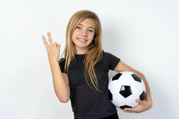 ホワイトウォールの上にサッカーボールを保持しているスポーツウェアを身に着けているティーンガールは笑顔でフレンドリーに見え 前方に手でナンバー2または秒を示し カウントダウン — ストック写真