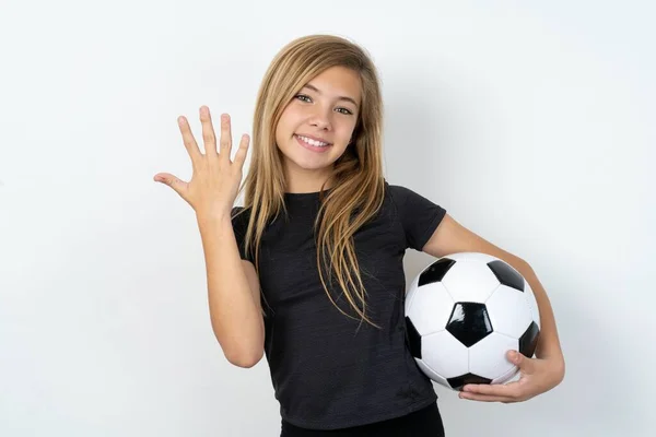 ホワイトウォールの上にサッカーボールを保持し 笑顔でフレンドリーに見えるスポーツウェアを着ている10代の少女は 前方に5番目または5番目の数字を示し カウントダウン — ストック写真