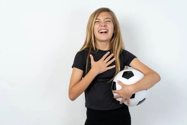 白い壁の上にサッカーボールを保持するスポーツウェアを身に着けている美しい白人十代の少女は幸せを表現し 喜んで笑い 心に手を置きます — ストック写真
