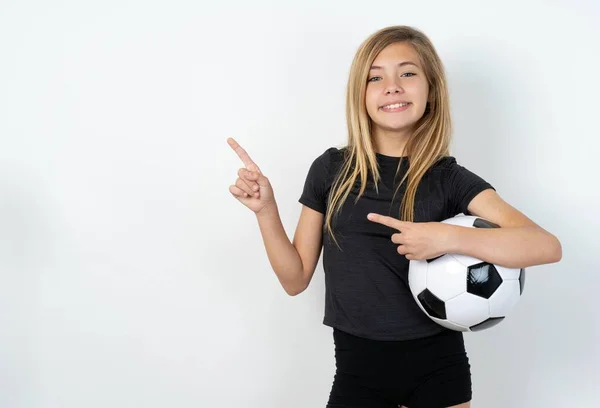 コピースペースでホワイトウォールポイントの上にサッカーボールを保持するスポーツウェアを着ているティーンガールは 広告が正しい方向を与えることを示しています — ストック写真