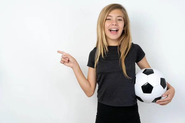 身穿运动服的少女拿着足球在白墙上欢快地笑了起来 指出空旷的空间展示了购物打折的提议 好消息或意想不到的销售让她兴奋不已 — 图库照片