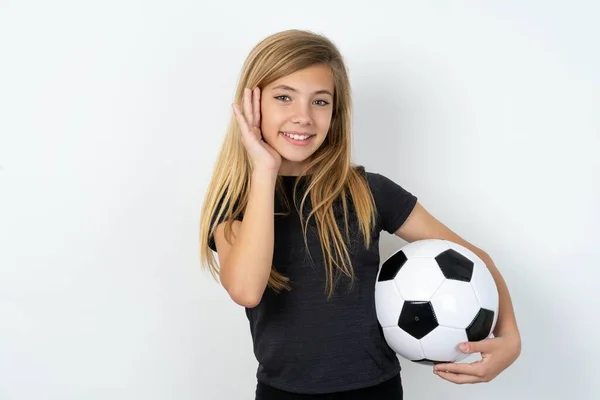 快乐的少女穿着运动服 拿着足球从白墙上拿过 温柔地触摸着两颊 面带微笑 露出洁白的牙齿 积极地凝视着镜头 — 图库照片