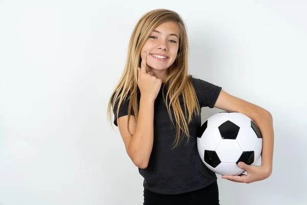 ハッピーティーンガールは 歯痛のある笑顔でホワイトウォールの上にサッカーボールを保持するスポーツウェアを着用し 口の近くに指の指を保持し 陽気な笑顔を強制します — ストック写真