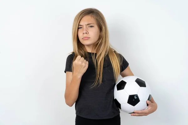 身穿运动服的少女拿着足球在白墙上飞舞 表示拳头惹怒了脸上的表情 要报复或威胁要有人严肃地看 我让你看看谁是老大 — 图库照片