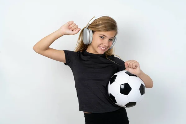 穿着运动服的无忧无虑的少女 带着足球在白墙上飞舞 面带微笑 提起臂膀 舞姿悠闲 乐声悠扬 通过耳机聆听着演奏者的音乐 — 图库照片