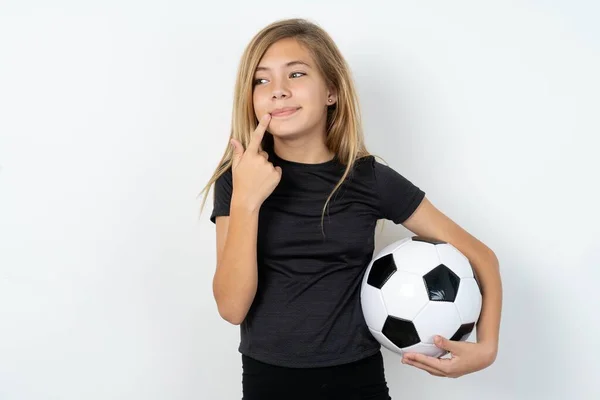 白い壁の上にサッカーボールを保持するスポーツウェアを身に着けている美しい夢のようなティーンガールは 唇の近くに指を保ちます コピースペース — ストック写真