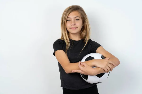 自信を持って十代の少女は カメラを見ている腕と白い壁の上にサッカーボールを保持するスポーツウェアを着ています — ストック写真
