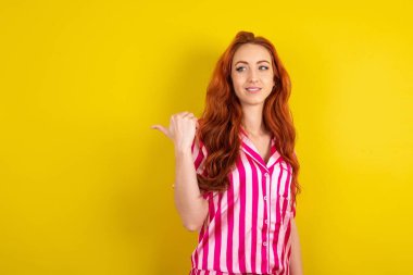 Sarı stüdyo arka planında pembe pijama giyen genç kızıl saçlı kadın fotokopi alanında reklam konseptini gösteriyor.