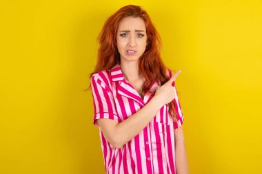 Sarı stüdyo arka planında pembe pijama giyen genç kızıl saçlı kadın işaret parmağı, endişe ve sürpriz konsepti bir yana bırakırsak endişeli ve gergin..