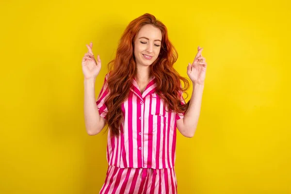 黄色いスタジオの背景にピンクのピンクのパジャマを着用している若い赤毛の女性は大きな希望を持っています 指を交差 幸運を信じています 広く笑顔 人と願いの概念 — ストック写真
