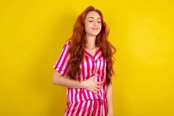 黄色いスタジオの背景にピンクのピンクのパジャマを着用している若い赤毛の女性は 穏やかに微笑み 食べて満足の概念に触れます — ストック写真