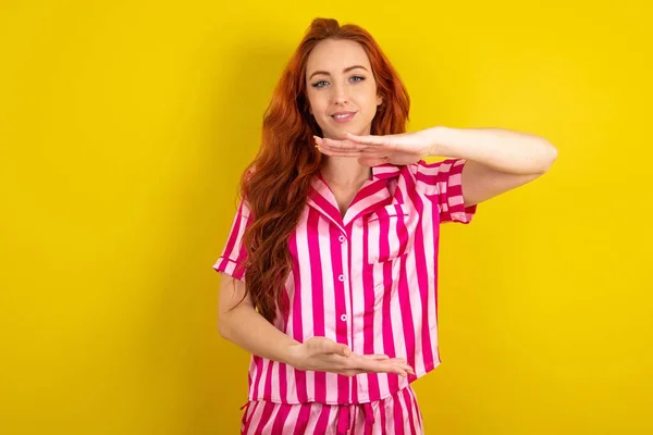 黄色いスタジオの背景にピンクのピンクのパジャマを身に着けている若い赤毛の女性は 大きくて大きい看板を示す手でジェスチャーし シンボルを測定します カメラを見ている笑顔 — ストック写真